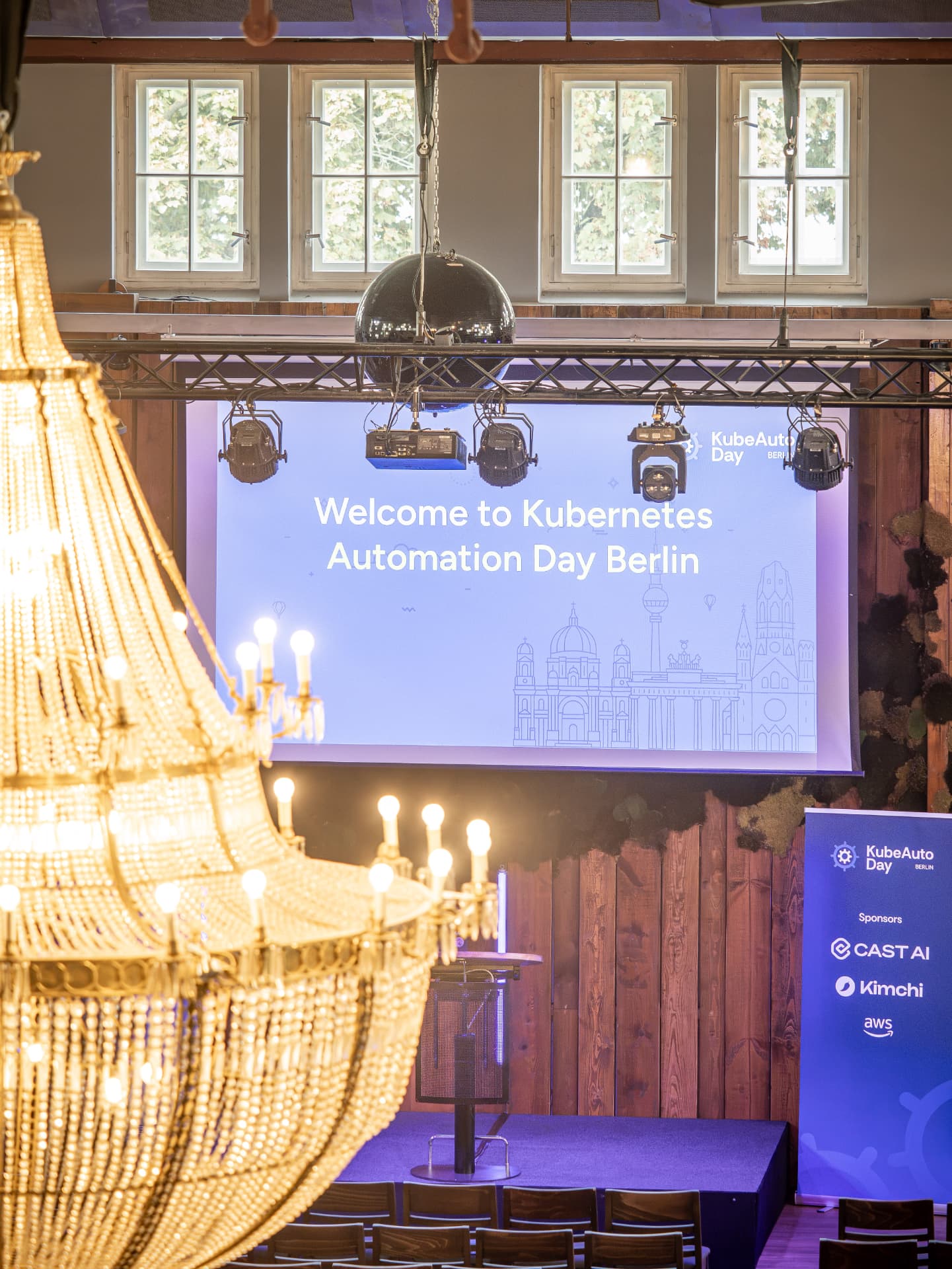 Welcome screen for KubeAuto Day Berlin above the stage, framed by a chandelier